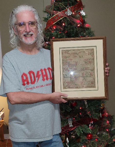 author holding a framed cross-stitch sampler piece from 1832