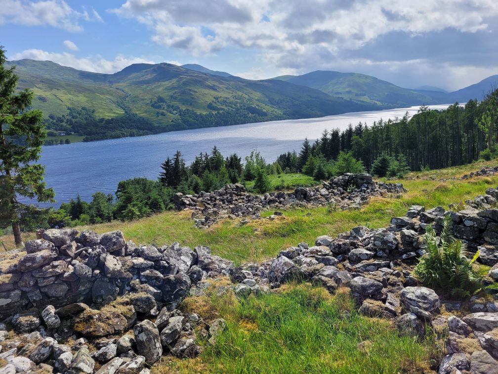 foreground - ruins of the foundation of an 18th century Highland Scottish longhouse and the slope of a mountain looking down onto a loch, taken on a sunny summer day. The floor is overgrown with long grass. The ruins are surrounded by grass with a pine forest in the distance on the slope down to the lochside. It is a bright sunny day with scattered, fluffy white clouds. The lake is sparkling from the sunlight.